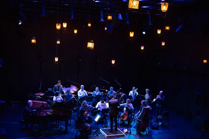 An ensemble of musicians performs on stage under hanging lamp light.