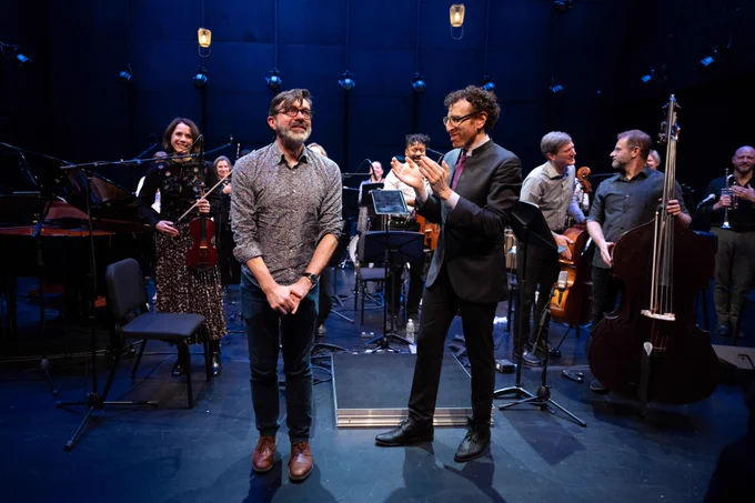 Two people, Donnacha Dennehy and Alan Pierson stand together in front of Alarm Will Sound on stage.