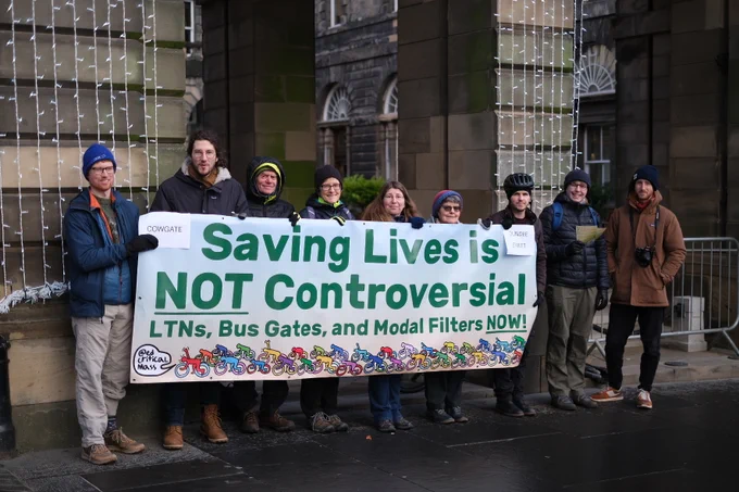 A group of people wearing winter clothes standing in front of the Edinburgh City Chambers holding a banner that says "Saving lives is not controversial! LTNs, bus gates and model filters now!"
