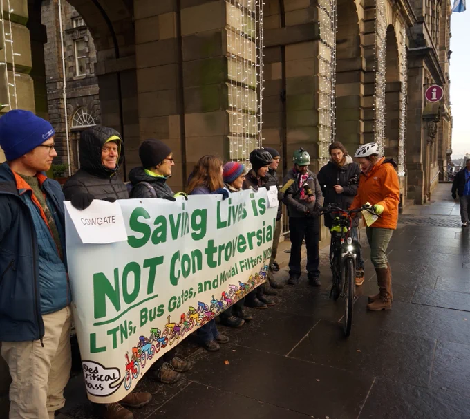 A group of people wearing winter clothes holding a banner that says "Saving lives is not controversial!" Some of them are talking to Councillor Dijkstra-Downie, who is holding a bike and dressed in a bright orange coat.
