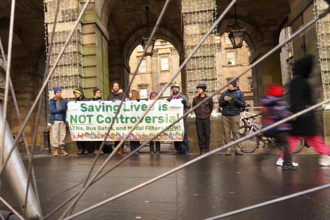 A group of people wearing winter clothes standing in front of the Edinburgh City Chambers holding a banner. In the foreground are the spokes of. The banner says "Saving lives is not controversial! LTNs, bus gates and model filters now!"