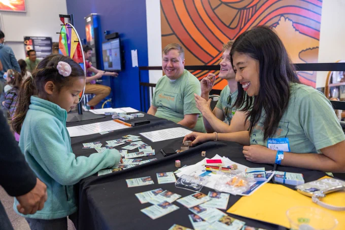A kid looking at the STEM trading cards on the table with three staff members smiling at her.