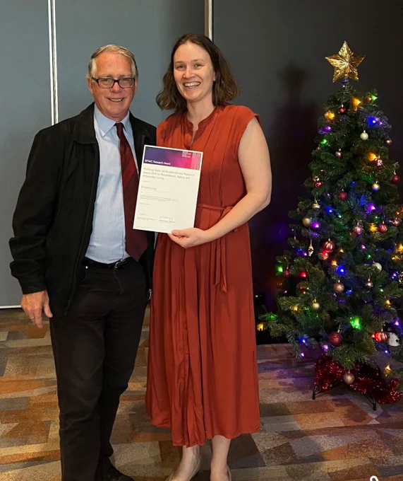 Two people standing in front of a Christmas tree, smiling at the camera. One person is holding an award certificate.
