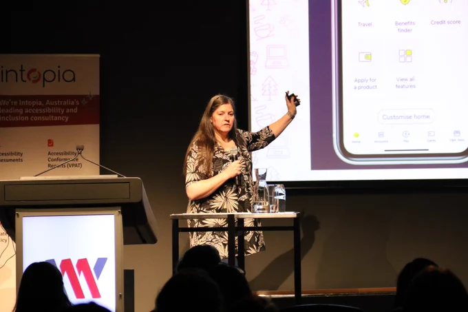 Allison Ravenhall onstage at A11y Camp pointing to the big screen behind her, which shows the CommBank app.