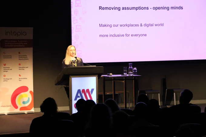Donna Purcell presenting at the lectern on stage at A11y Camp. The text on screen behind her "Removing assumptions – opening minds. Making our workplaces & digital world inclusive for everyone".