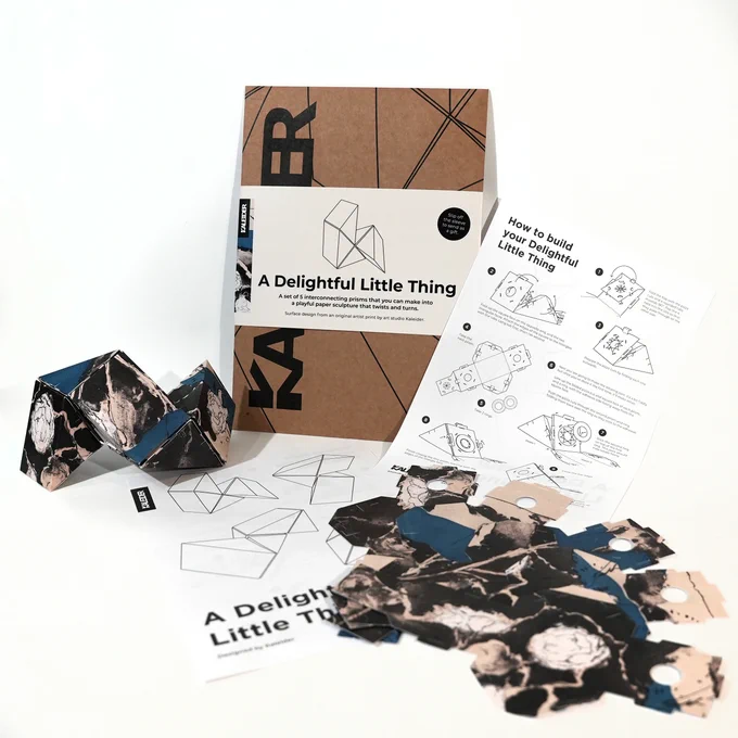 A product shot of Kaleider's Delightful Little Things against a white background. It shows a cardboard envelope package, a sheet of instructions printed on both sides, a set of five flat cardboard cutouts before assembling, and an assembled set. They are printed with a cream, black, and blue rose design.
