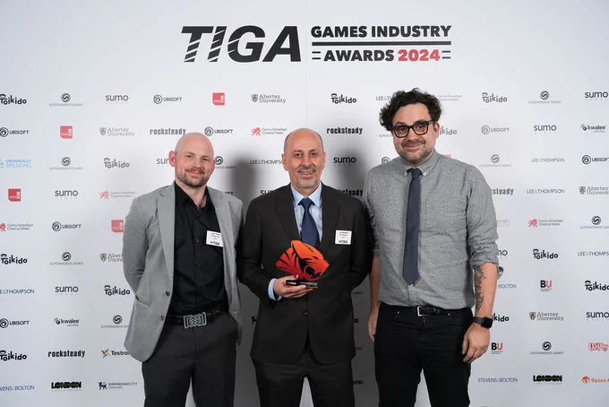 Three game developers hold a TIGA video game trophy in front of a banner that says TIGA Games Industry Awards 2024
