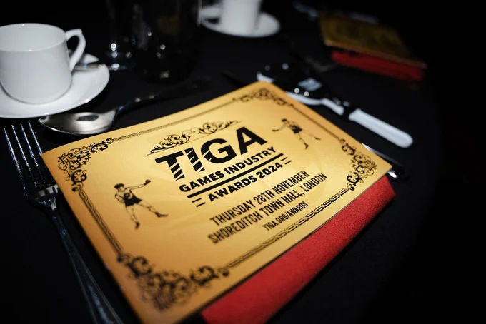 A gold pamphlet lies on a table. The cover says TIGA Awards 2024, Thursday 28th November 2024, Shoreditch Town Hall, London