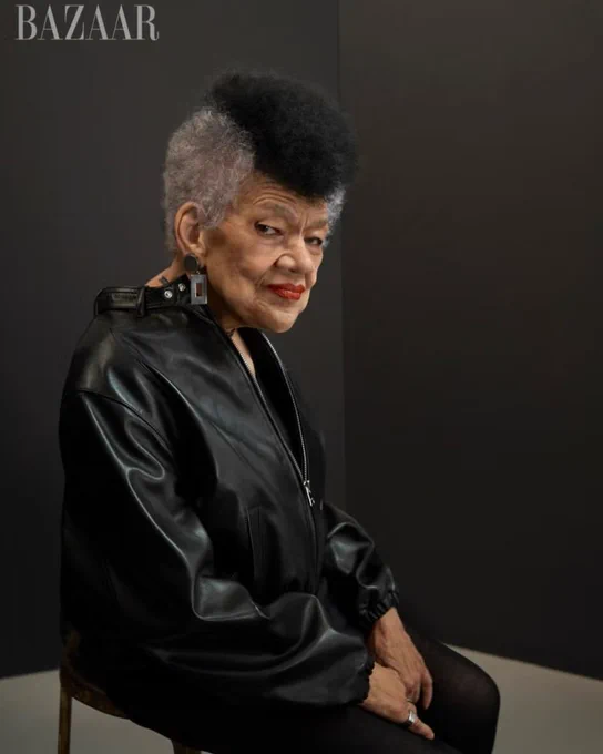 Lorraine O’Grady, a Black woman with salt and pepper hair, sits for a portrait in a black leather jacket, black tights, long earrings, and red lipstick.