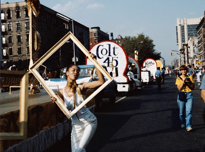 A Black woman in a white dress holds a large  gold frame while standing in front of a parade float during Harlem’s African-American Day Parade September
1983.