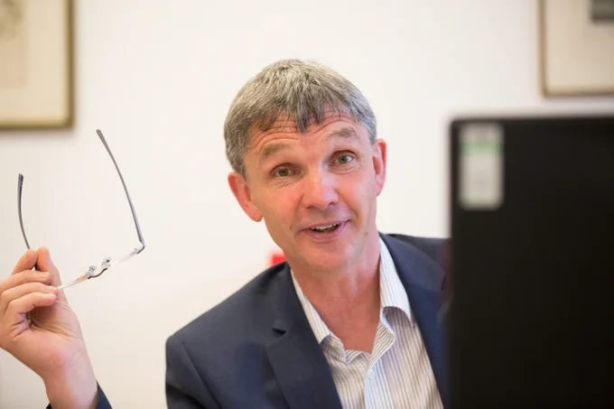A photo of Chris Gledhill sat behind a monitor smiling at the camera while holding his glasses