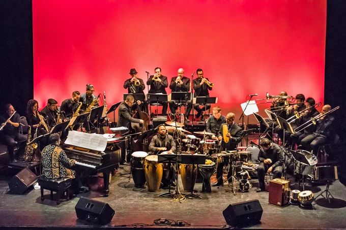 A large musical ensemble featuring people playing the piano, brass, and percussion instruments on stage in front of a red wall.