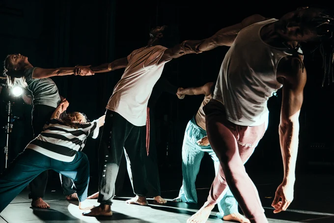 People dressed in street clothing dance on stage, holding each other by their outstretched arms.