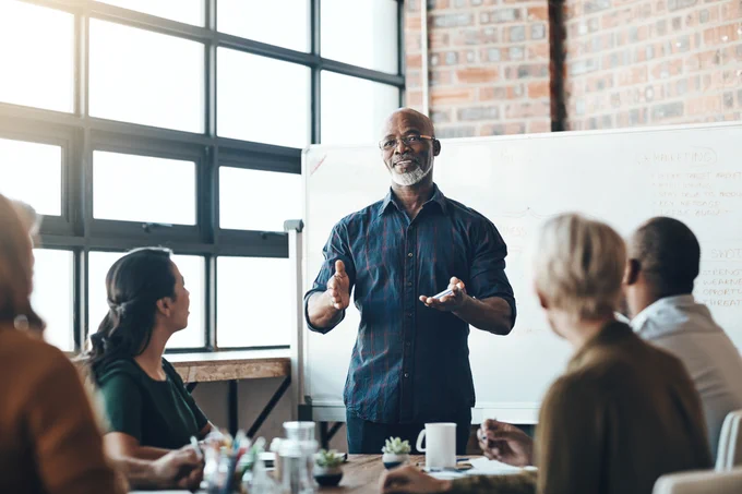 Stock image of a group of people sitting. A man is standing in front of a dry erase board, holding out both hands as others seated at the table listen in.