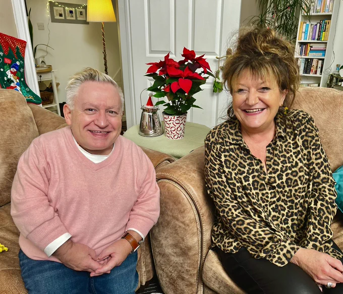 Simon on the left in a pink soft jumper and white t underneath. Jane is a blue tiger print blouse. Both sat on sofas with a poinsettia in the middle.