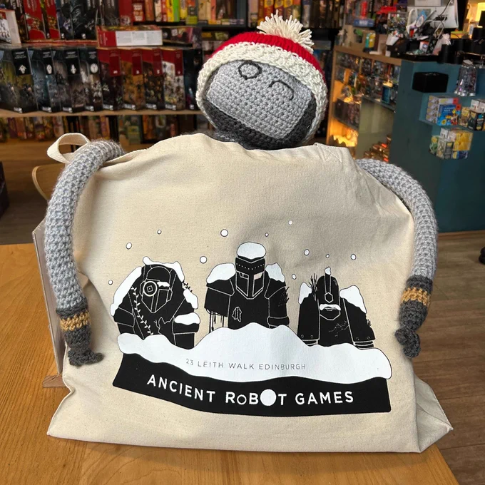 A Christmas tote bag with three robots, being held by a crocheted robot in the Edinburgh board game shop Ancient Robot Games.