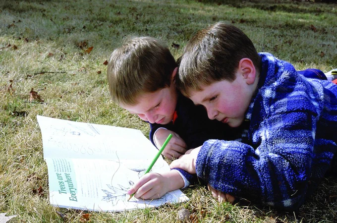 Two boys lay in the grass next to each other looking at a coloring book. The boy farthest away is drawing in it with a colored pencil.