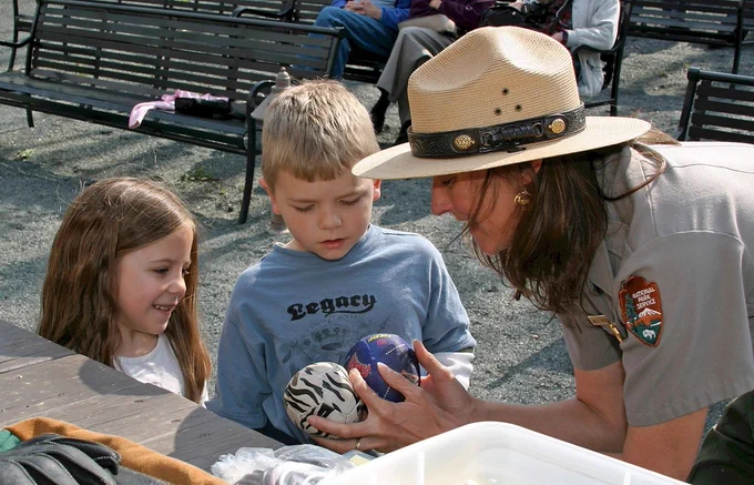 Two youth stand next to a woman wearing a park ranger uniform and hat who is holding two small balls in front of them