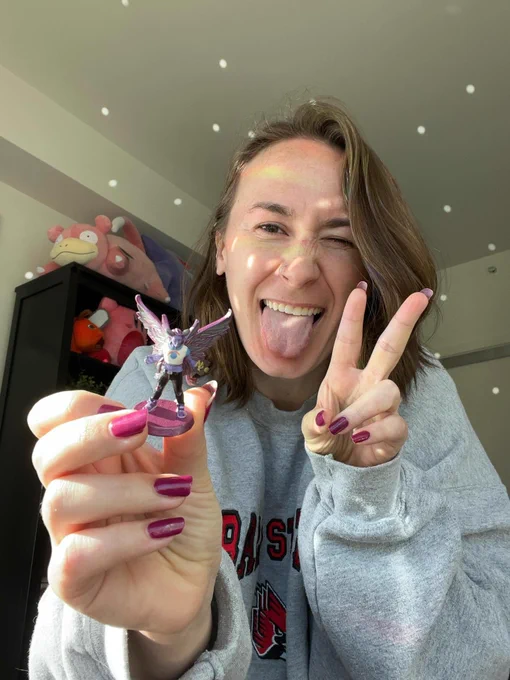 a photo of jes holding a mini of their character who is a moth person with wings. Jes is holding up a peace sign with their other hand and is sticking their tongue out