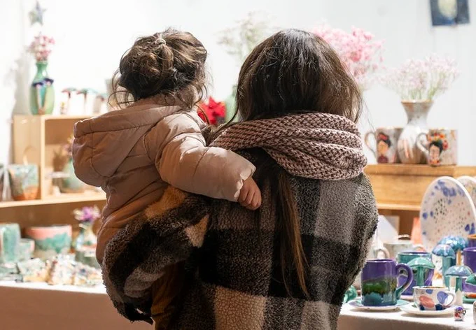 Una noia porta en braços una nena. Van abrigades, i estan mirant una paradeta, que sembla d'un mercat de Nadal.