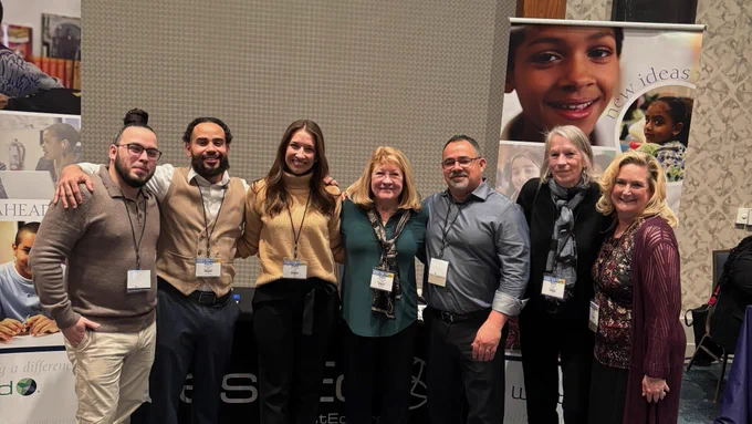 Group of seven staff members smiling and posing together at the WestEd conference booth with banners featuring children and youth. 