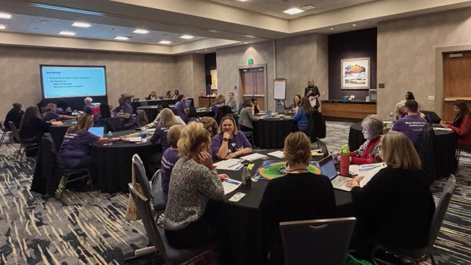 A group of attendees at a conference sitting at round tables in a large room with a presentation screen displaying text. Several individuals are engaged in conversation. 