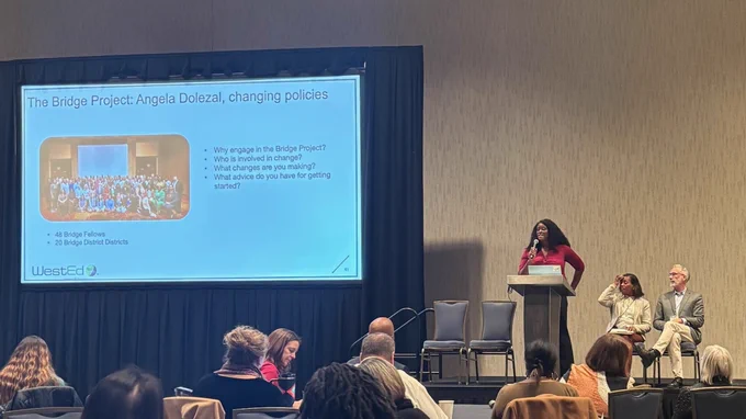A presentation at a conference with three panelists seated and one standing next to a large screen. The audience is attentively watching the presentation.