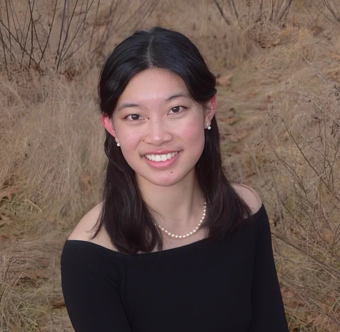 Portrait of a smiling person with shoulder-length hair, wearing a black top and a pearl necklace, set against a background of dry grass and bare branches.