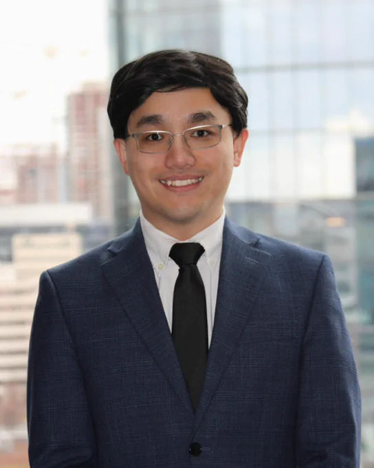 Person in a blue suit and black tie standing in front of a window overlooking a city skyline.