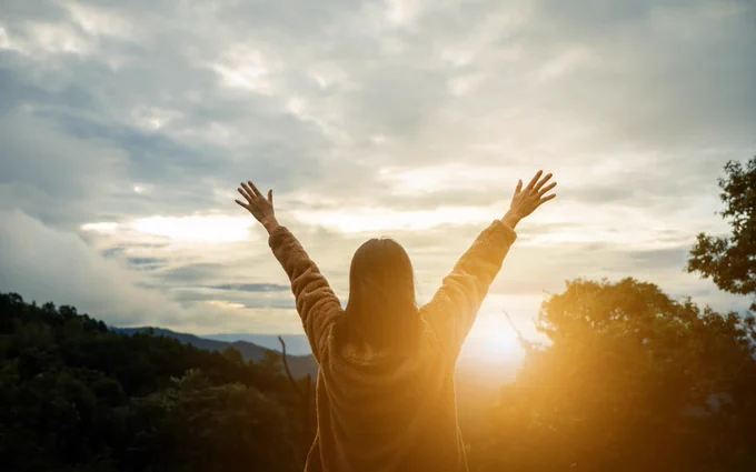 A person stands with arms outstretched toward the evening sun