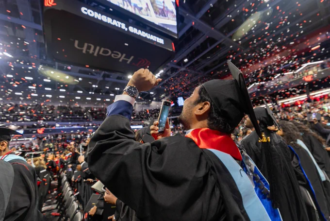 Graduating student in cap and gown celebrating at commencement ceremony, raising a fist triumphantly amid falling confetti, with 'CONGRATS, GRADUATE!' displayed on a large screen overhead.