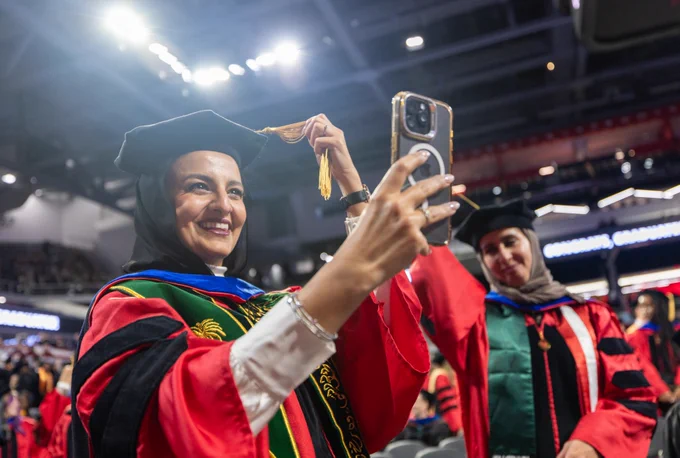 Graduate in a black cap and decorative red and black gown taking a selfie with a smartphone at a graduation ceremony, with another graduate in the background.