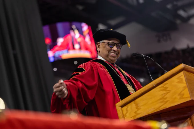 President Pinto in academic regalia delivering a speech at a university graduation ceremony, with a large audience in the background and a projection screen visible above.