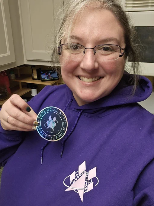 A person smiling while holding a circular sticker with the text "Intergalactic Space Beans" and "Meow" on it. They are wearing glasses and a purple hoodie with a matching logo of a stylized rocket and planet design on the chest. The background shows a kitchen area.