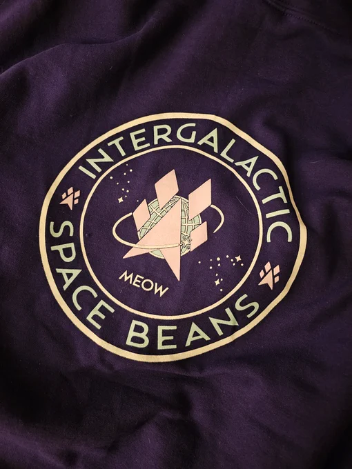 A close-up of a purple fabric featuring a circular logo with the text "Intergalactic Space Beans" and "Meow." The design includes a stylized rocket surrounded by stars and planets, emphasizing a space theme.