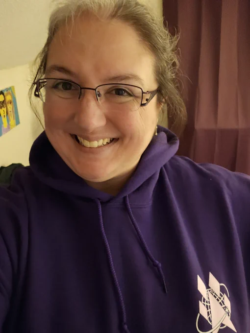 A person smiling while wearing glasses and a purple hoodie with a logo on the chest featuring a stylized rocket and planetary design. The background includes a piece of colorful artwork and a curtain