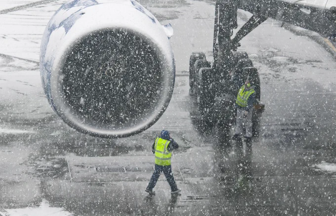 Ground crew in high-visibility jackets working near an aircraft during a snowfall at an airport.