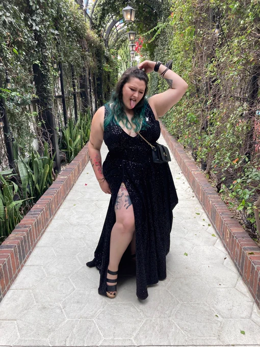 A photo of me, an average height white girl with teal hair in a floor length black sequin gown with slits up both sides. My leg is out in front and the dress has a deep v. I am sticking out my tongue. Around me is an arched tunnel covered in climbing vines 