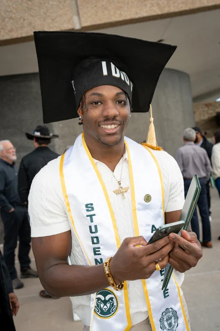 A C S U graduate wears an oversized graduation cap and smiles for a photo outside of Moby Arena.