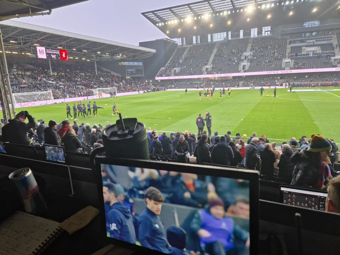 Photo from media area showing a live screen of the match, the pitch with players warming up and putney and riversides stands slowly filling up