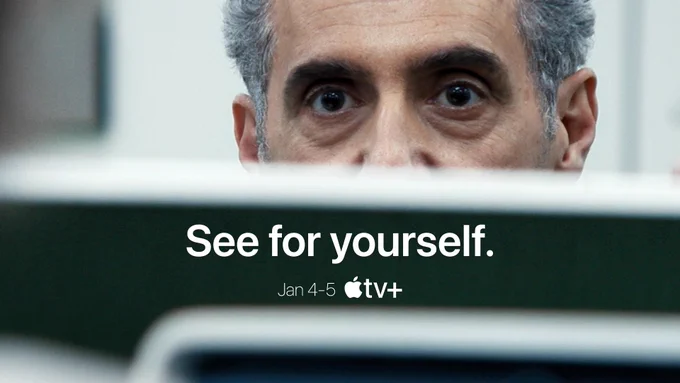 From Severance. A close-up shot of John Turturro as Irving reveals his eyes peering blankly over a divider at Adam Scott as Mark, The bottom of his face is hidden by the divider. Text on the image reads, “See for yourself. Jan 4 and 5.” The Apple TV  logo is displayed next to the text.