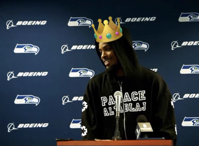 Smiling Devon Witherspoon is wearing a black hoodie with the hood up as he stands at the post-game presser lectern mic. The backdrop is navy with the Seahawks and Boeing logos. There is an emoji crown added to his head as he looks to the left.