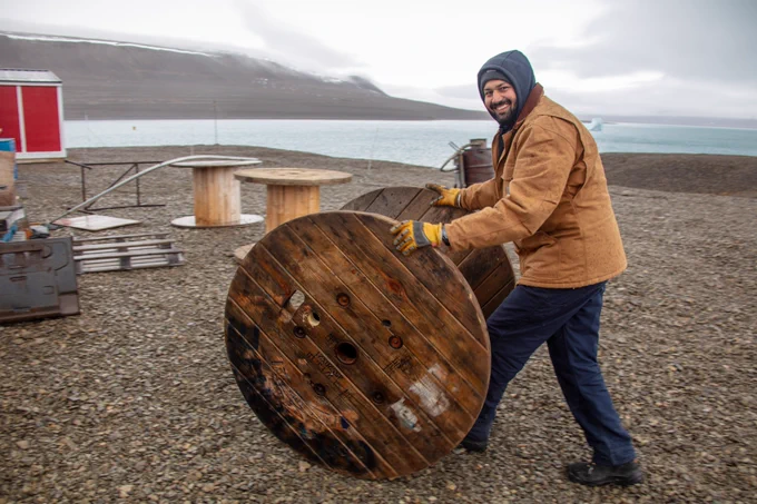 Person in beige jacket and blue jeans rolling a large round wooden object on pebbled ground. Some metal equipment is on the left; water and rocky land is behind in the distance.