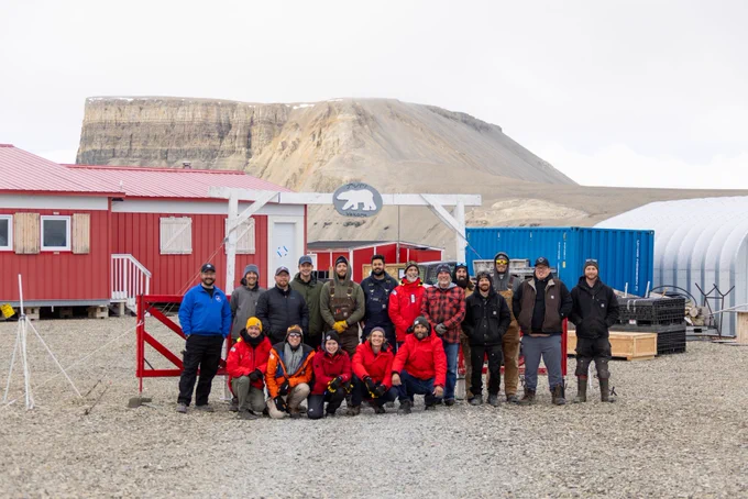 Group of 16 people standing together in jackets on pebbled ground. Red building with white trim to the left, and equipment in the background. Rockland in distance behind group of people.