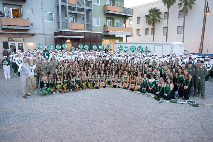 Group photo at the Arizona Bowl pep rally featuring the C S U marching band, cheer team, poms team, color guard, ROTC members, Cam the Ram mascot, President Amy Parsons, and the athletic director. The group is gathered in front of the M L K Apartments, with Rams logos displayed on signs and equipment in the background. The cheer and poms teams are in the front holding green and yellow pom-poms, with the band and other participants standing behind them. Everyone is smiling, creating an energetic and spirited atmosphere.