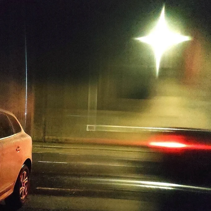 En una calle de noche vemos parte de un coche blanco aparcado a la izquierda. El trazo de la luz trasera de color rojo de un coche negro a mano derecha. Una luz brillante en forma de cruz arriba del coche negro