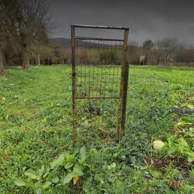 Puerta solitaria formada por reja de hierro en un campo verde. Es una puerta sin uso, ya que no hay vallas. Cielo con nubarrones.
