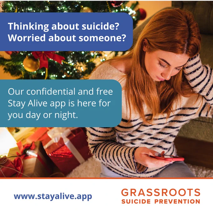 Text reads 'Thinking about suicide? Worried about someone?', 'Our confidential and free Stay Alive app is here for you day or night.' Photo shows someone looking at their phone while sitting next to a Christmas tree where lights, baubles, and presents can be seen.
