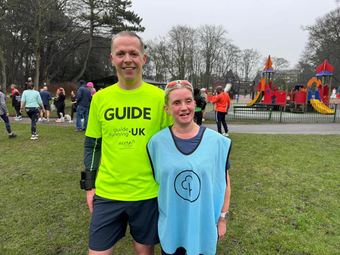 The image shows two people standing on a grassy area in a park. The person on the left is a man wearing a bright yellow-green shirt with the word "GUIDE" in large black letters on the front. Below "GUIDE," the shirt says "guide running UK" and "acorns putting people first." He is also wearing dark shorts and has a black armband on his left arm. The person on the right is a woman wearing a light blue vest with a circular  logo. She is smiling and has sunglasses resting on her head. In the background, there are several other people, some walking and some standing, and a colorful children's playground with slides and climbing structures. The sky is overcast, and there are trees without leaves surrounding the area.