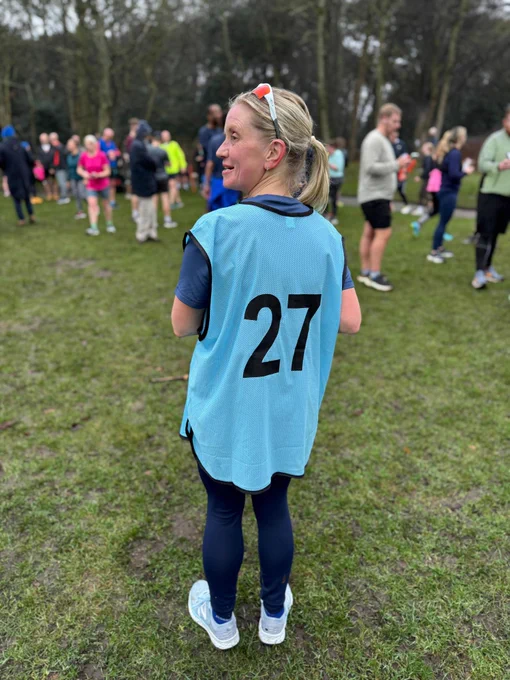 The image shows a woman standing outdoors on a grassy area, possibly a park or field, with a group of people in the background. She is wearing a light blue vest with the number 27 printed on the back in black. She has blonde hair tied back and is wearing sunglasses on her head. Her outfit includes dark blue leggings and light blue running shoes. The people in the background are dressed in athletic clothing, suggesting they might be participating in a group event or activity, such as a run or sports event. The setting appears to be a cloudy day with trees in the background.